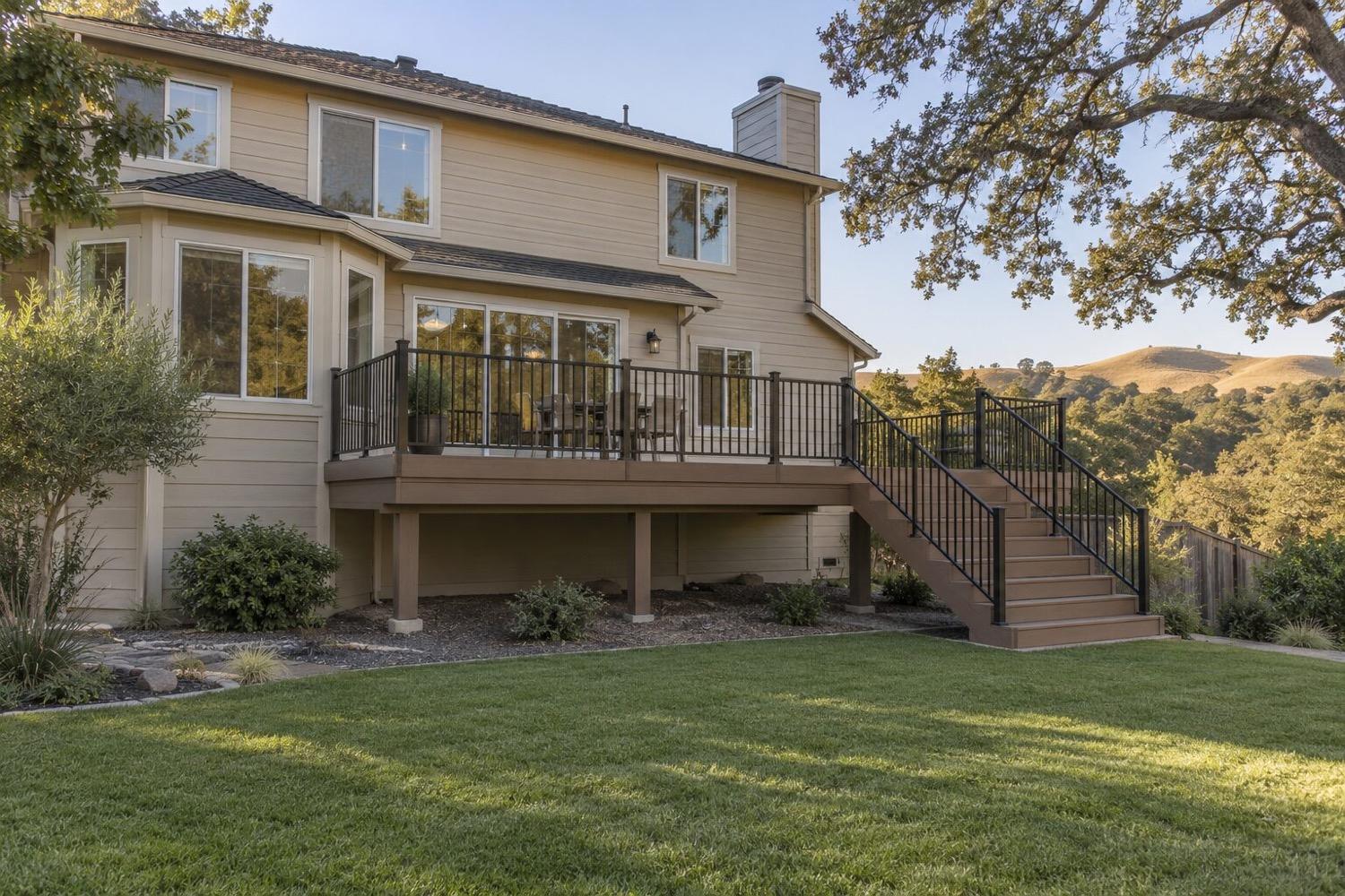 Raised brown composite deck with black aluminum railings and a wide stair down to the lawn, across the back of a tan two-story home with oak-covered hills behind.