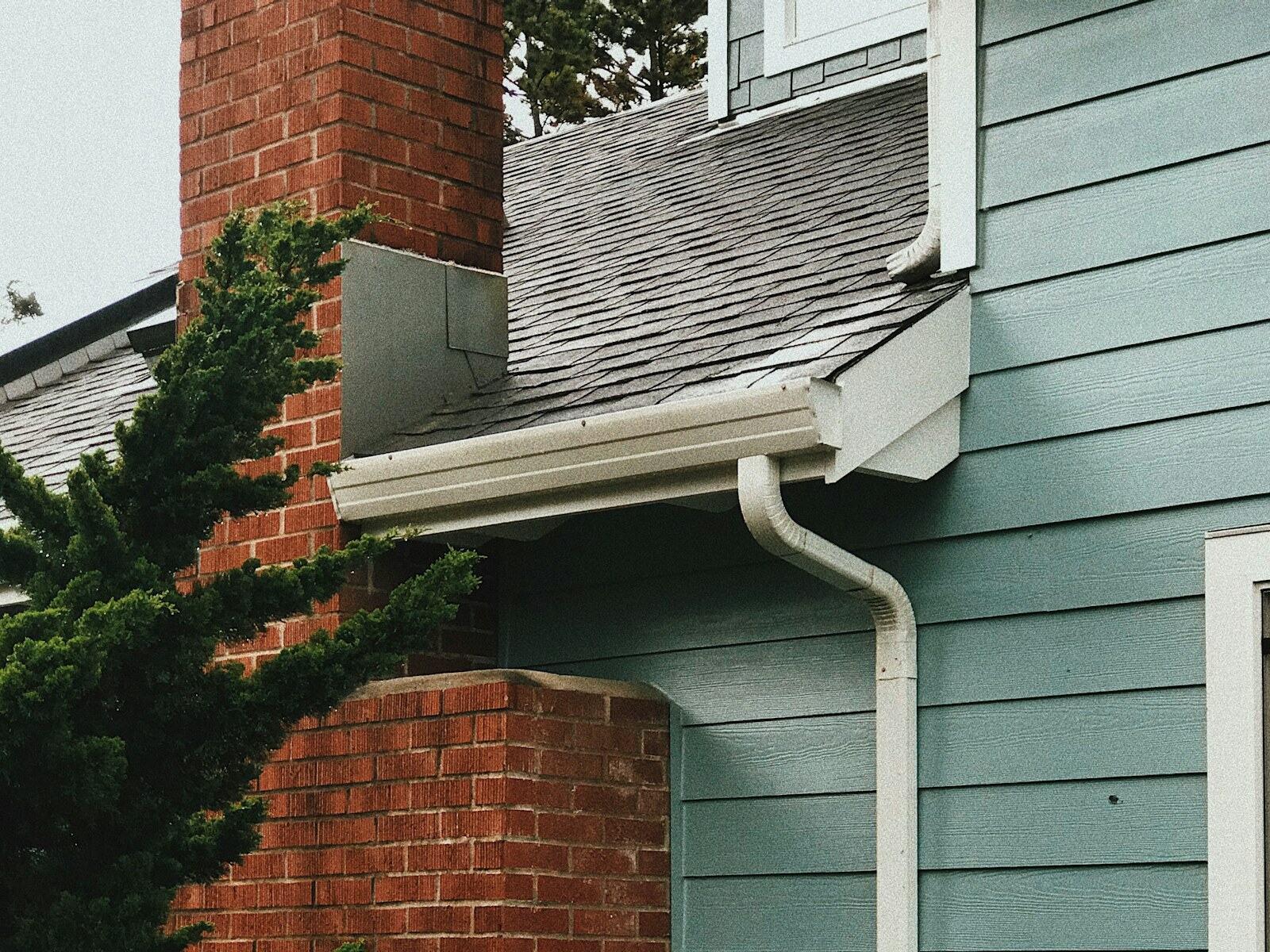 Downspout and gutter beside a brick chimney on gray-green siding.