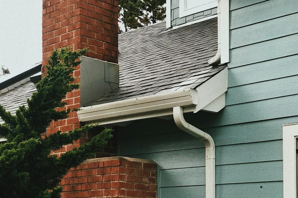 Downspout and gutter running past a brick chimney on gray-green siding.